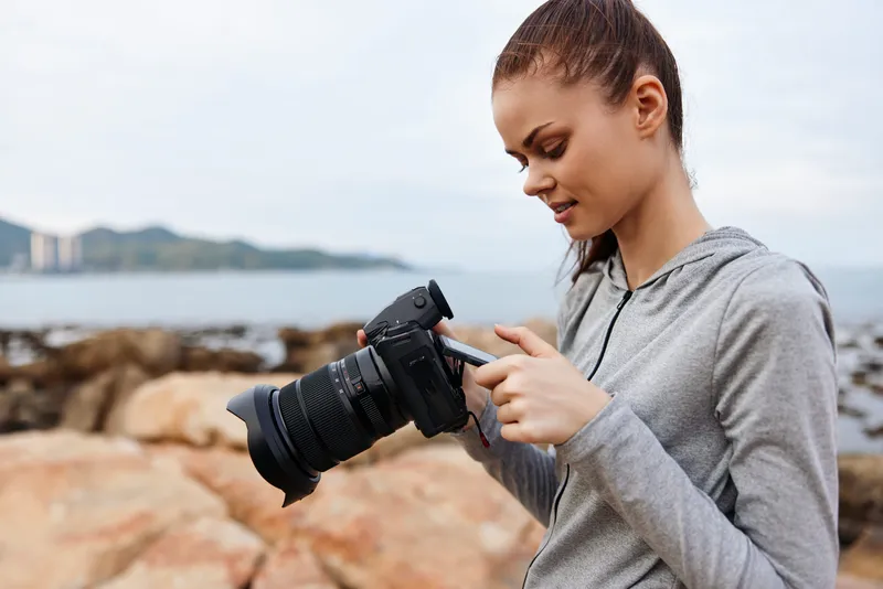 Young female photographer in casual gray hoodie enjoys taking photos with a professional camera by the seaside, capturing the beauty of nature and her passion for photography