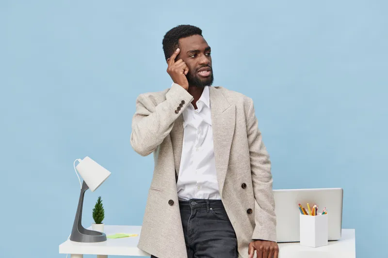 Thoughtful man in beige blazer and white shirt, standing at modern office desk with computer and stationery, looking away with hand on temple, indoor workspace background, business and professional co