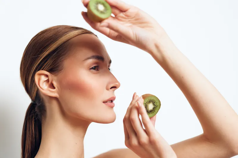 Woman kiwi skincare face fruit beauty mask wellness concept with a young adult applying kiwi slices to her cheek for natural hydration and glowing skin in a clean bright studio