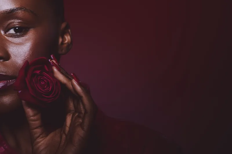 Close-up of a dark skinned African woman holding a red rose near her face. The image features elegant beauty, floral concept, and natural skin texture on a dark background.