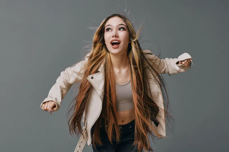 Young woman with long braided hair wearing a light jacket and crop top posing energetically against plain background. Expression shows excitement and movement in studio.