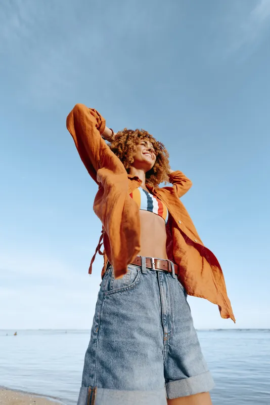 Young woman with curly hair enjoys a sunny day at the beach wearing a striped crop top and an orange open shirt with denim shorts, feeling relaxed and carefree under the clear blue sky.