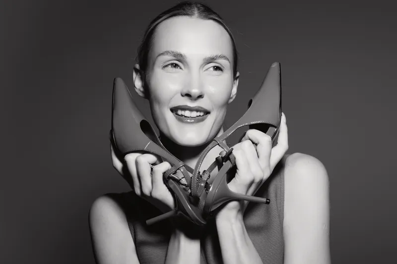 Smiling Caucasian woman with fair skin holds high heel shoes near face, showing joyful emotion and fashion concept in monochrome studio photo.