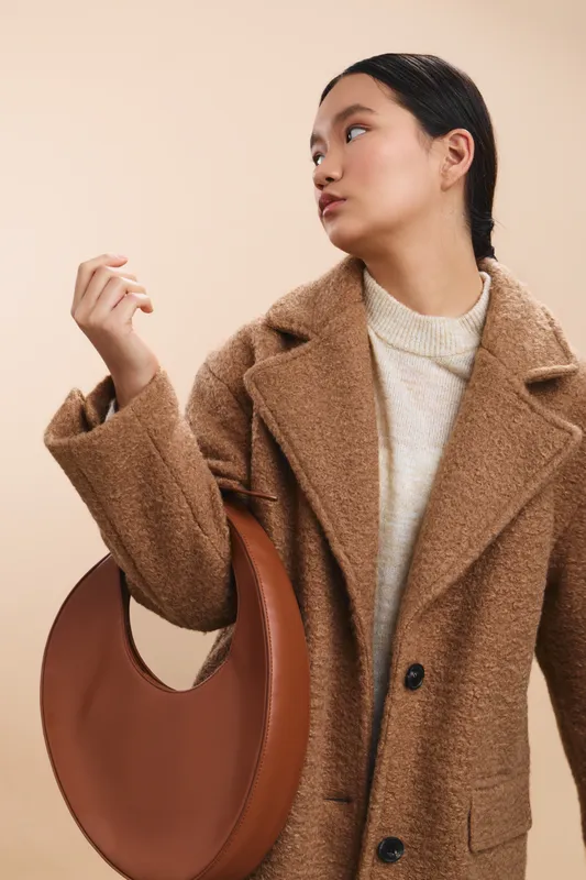 Asian woman in a chic tan coat stands against a warm beige backdrop, glancing to the side while holding a circular brown handbag, exuding modern style and confidence.