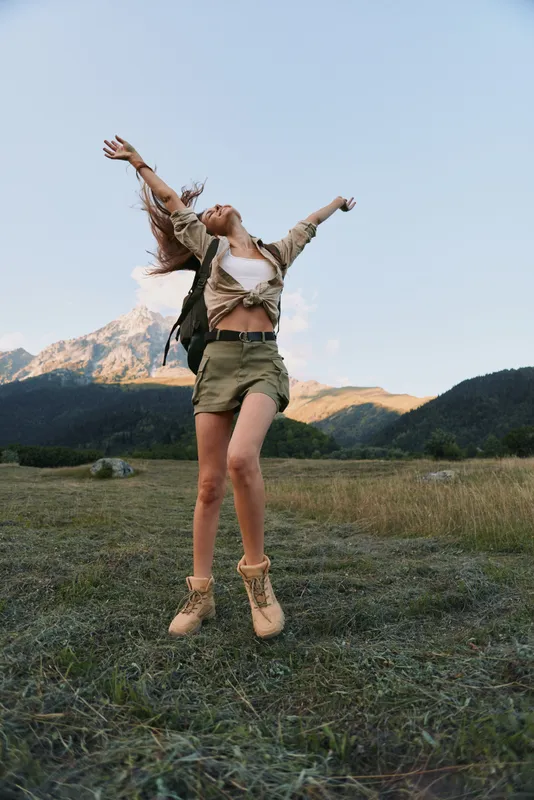 Joyful woman jumping in a grassy field with mountains in the background, depicting outdoor lifestyle, freedom, and carefree adventure under a clear sky.