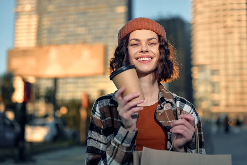 woman coffee shopping smile urban city portrait happy female enjoying takeaway cup and bag in sunlight with authenticity and candid lifestyle vibe, golden hour glow and mindful living emotional