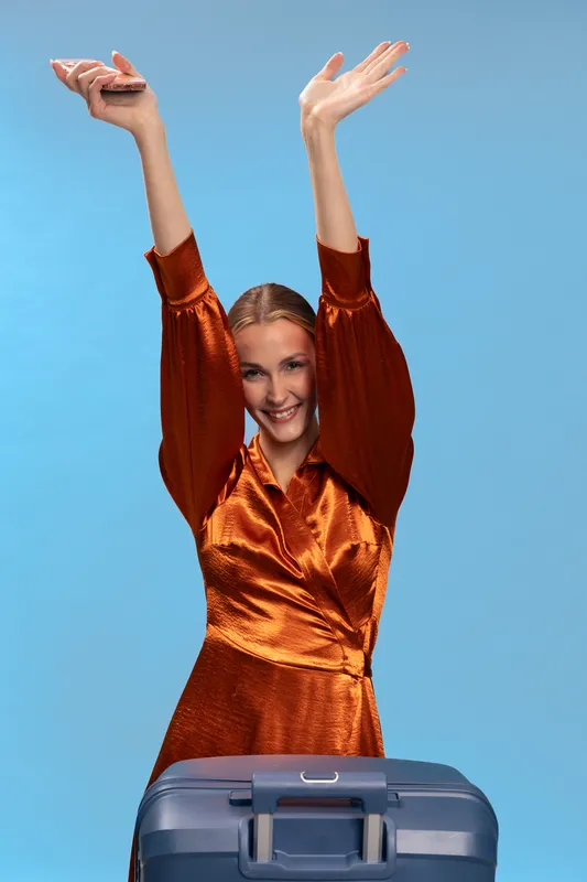 Happy woman in shiny orange dress raises hands up while standing behind suitcase. Smiling traveler ready for vacation or business trip on blue background. Concept of travel and joy.