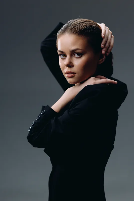 serious young woman with braided hair posing in black outfit with gray background, confident expression, studio portrait