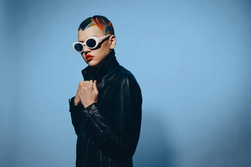 young woman with colorful hair and sunglasses wearing a black leather jacket against a blue background, showcasing a bold and edgy fashion style.