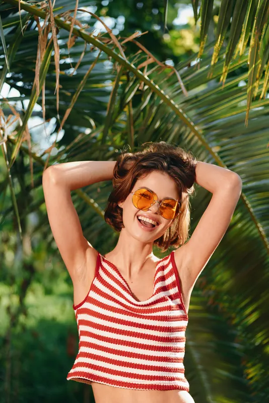 A cheerful young woman wearing a red striped crop top and round sunglasses smiles brightly while posing outdoors among palm fronds, enjoying sunny weather and a carefree vibe.