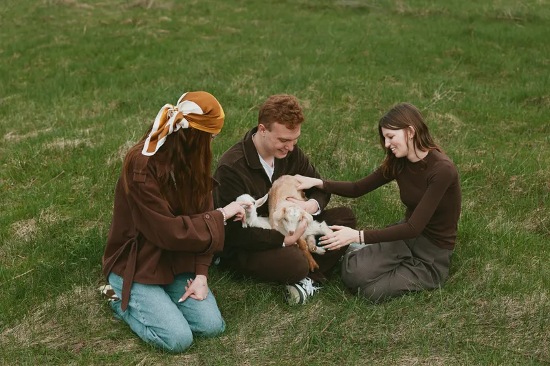 Two young women sit on grass in casual clothes while holding and petting a baby goat during a relaxed outdoor lifestyle moment in nature.