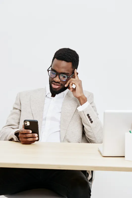 Confident smiling man of African descent wearing glasses and beige blazer sitting at desk with smartphone and laptop against plain white background, professional business casual look, lifestyle concep