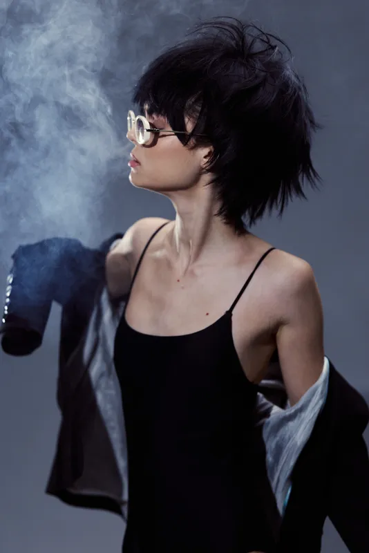 Stylish woman with short hair and glasses wearing black dress and smoking in dim light.