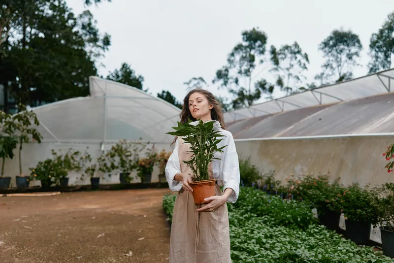 Young woman holding a potted plant in a botanical garden, showcasing joy and connection with nature in earthy tones and a serene environment