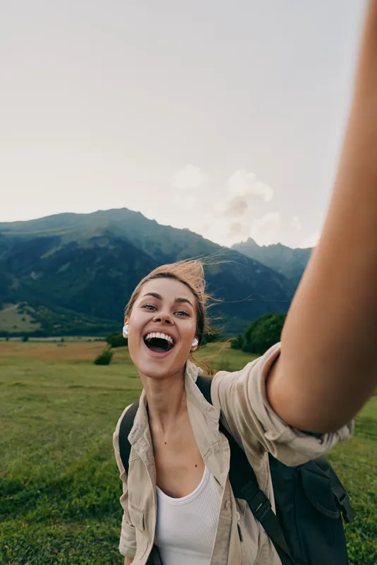 A joyful woman with a backpack takes a selfie in a grassy valley with distant mountains, radiating excitement and a sense of outdoor exploration.