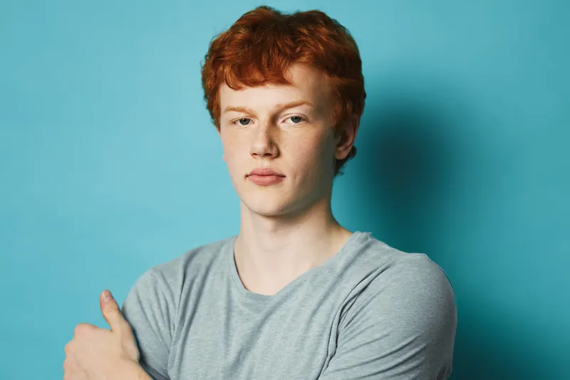 Young man with red hair and pale skin wearing grey t shirt stands against blue background looking straight at camera with serious facial expression and crossed arms.