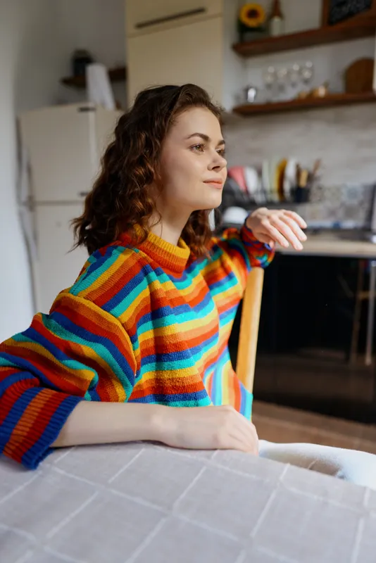 Cheerful woman at home, relaxed portrait in a colorful striped sweater, seated at a dining table, soft daylight, cozy kitchen backdrop, natural mood and casual lifestyle
