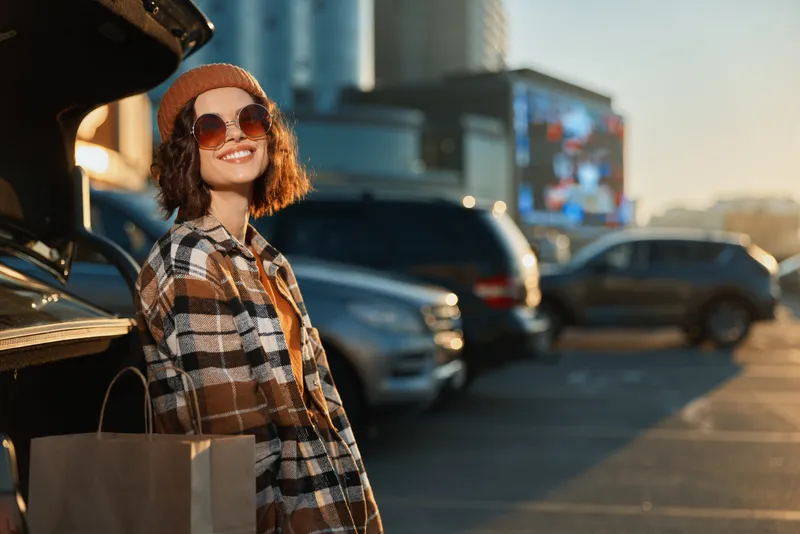 Woman shopping by car trunk with smile and sunglasses in urban parking, candid lifestyle moment with authenticity and golden hour glow, mindful living and emotional storytelling.