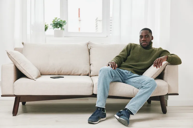 A relaxed young African man in a green sweater sits on a light sofa, showcasing casual home comfort and style in a bright space