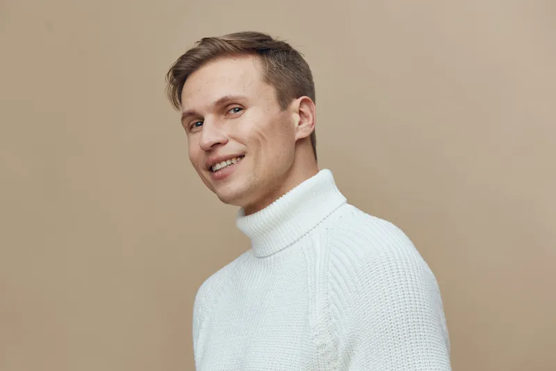 Smiling man young adult wearing white turtleneck sweater looking at camera studio portrait isolated on beige background cheerful expression lifestyle concept