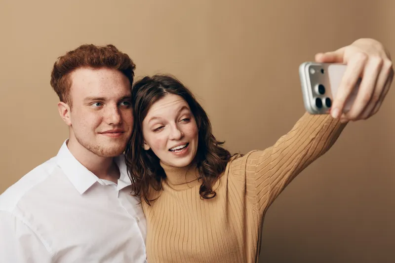 Young couple taking a selfie with a smartphone on beige background. The woman wears a brown sweater and smiles while the man looks at the camera. Concept of friendship and technology.