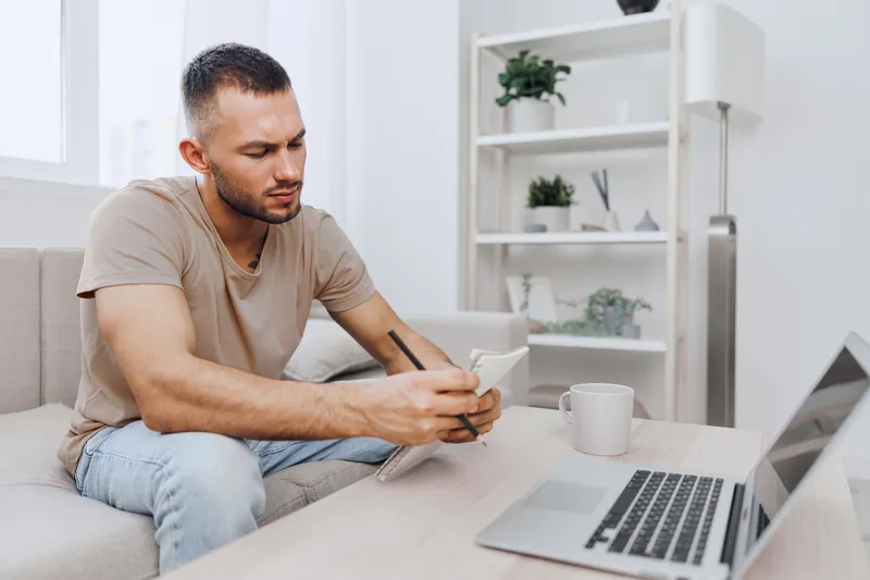 Thoughtful man taking notes at home office, dressed casually, engaged in creative process with a laptop nearby, plants in background, modern aesthetic.
