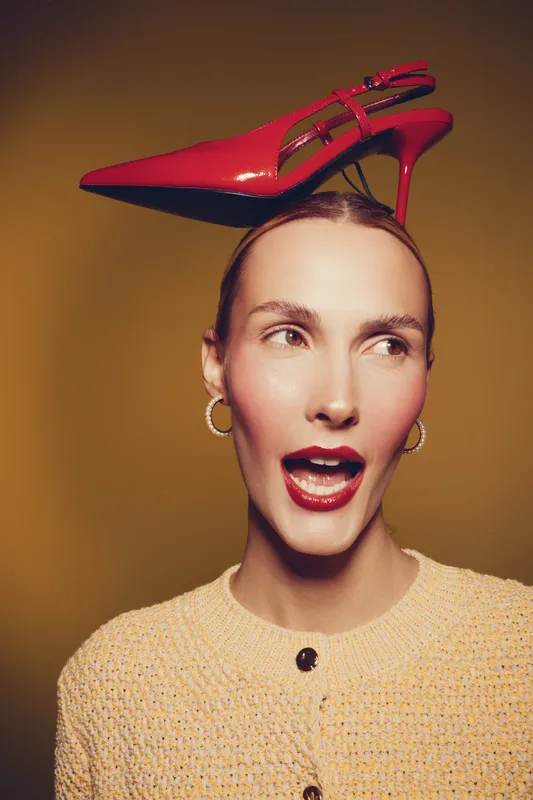 Young woman with red high heel shoe balanced on her head looks surprised. Studio portrait with warm lighting and yellow background. Concept of fashion, style, and playful attitude.