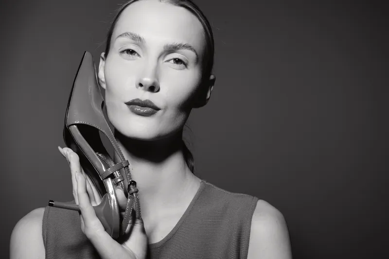 White woman with fair skin holds a high heel shoe near her face in black and white studio portrait. Concept of fashion, elegance, and minimalistic style with soft lighting.