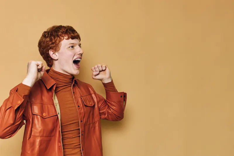 Young man with red hair celebrating success with clenched fists wearing brown jacket and turtleneck standing against plain background in studio.