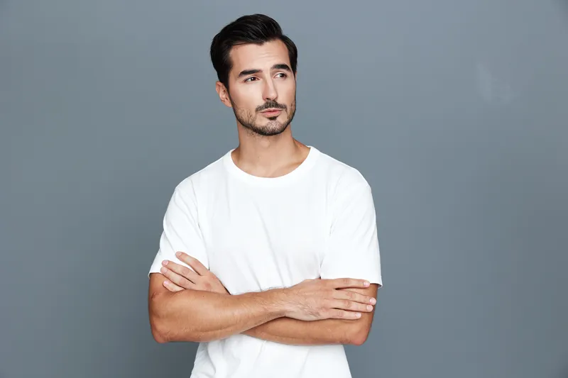 Handsome man with a thoughtful expression standing against a plain grey backdrop, crossing arms, dressed casually in a simple white t-shirt. Contemporary portrait concept.
