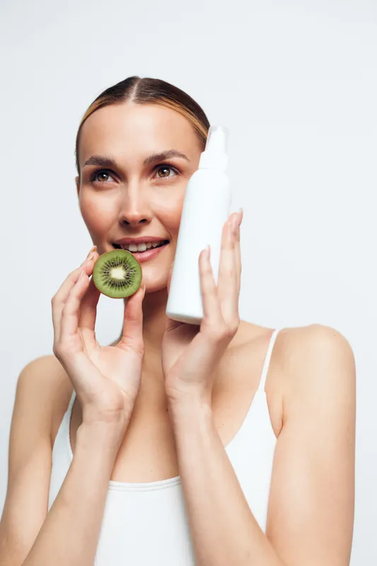 Woman skincare routine, holding kiwi slice and white lotion bottle, fresh face care, clean background, calm expression, natural look, studio lighting