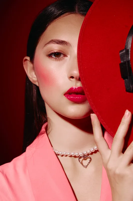 Young woman with glowy pink makeup and bright red lips hides half of her face behind a bold red hat. She wears a pearl necklace with a heart pendant. Beauty, fashion, creative concept.