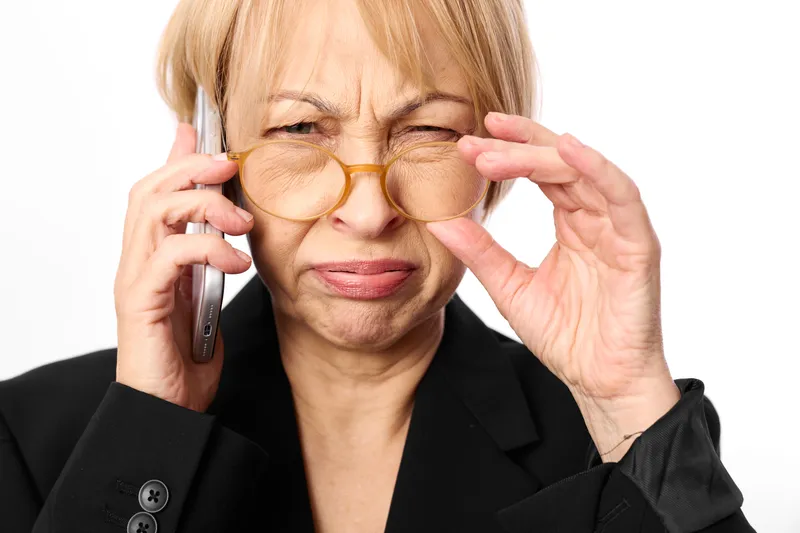 Elderly woman with glasses adjusting frames while speaking on smartphone phone call, business portrait isolated on white background, calm focused expression