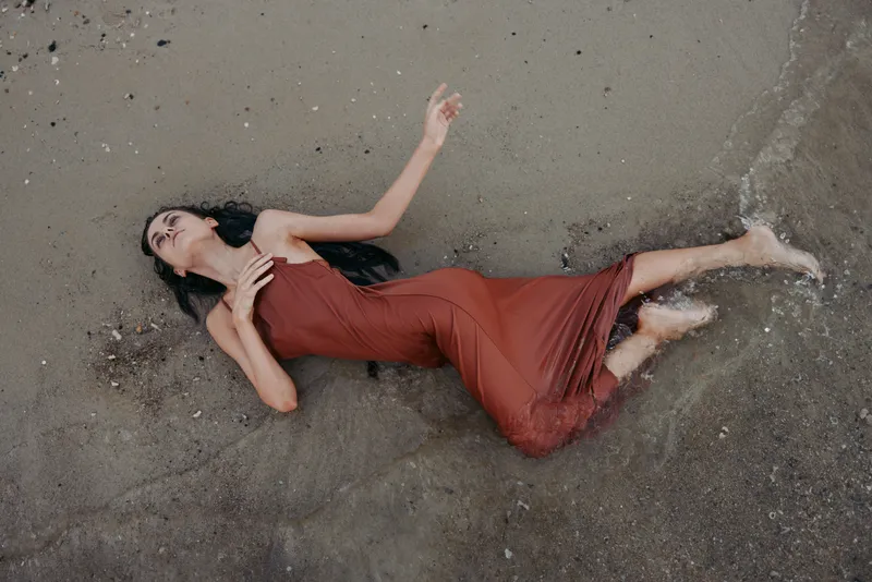 Portrait of a woman in a red dress lying on wet sand, arms raised and eyes closed, capturing vulnerability, motion, and a raw moment of surrender on a quiet shoreline.