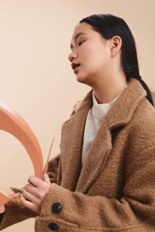 asian woman wearing a tan coat stands in a soft studio setting, holding a round accessory. she gazes slightly to the side with a calm, confident expression, conveying poise and focus.