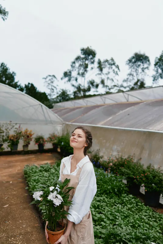 Young woman in an apron holding a potted plant in a greenhouse, showcasing happiness and connection with nature, perfect for seasonal gardening themes