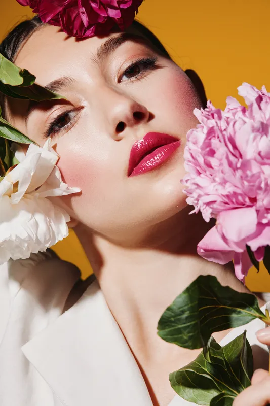 Close-up of fashion model with glossy red lips and floral adornments on face and hair. Bright orange background creates strong contrast with soft natural petals. Beauty creative concept.