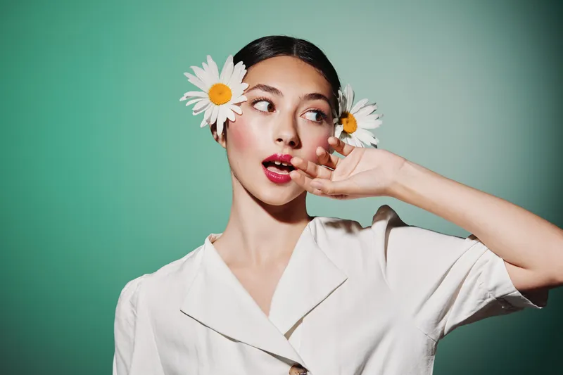 Young woman wearing white blouse poses with daisy flowers in her hair. She looks to the side with red lips and smooth skin. Bright green background enhances fresh beauty and fashion concept.