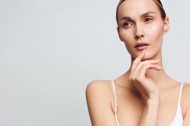 Thoughtful young woman with blonde hair and clear skin touches her chin while looking away against a light grey background in a minimalist skincare portrait.