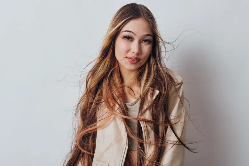 Young woman with long brown hair wearing light jacket looking at camera with soft smile on neutral background fashion, beauty, portrait and casual style with natural makeup and relaxed mood.