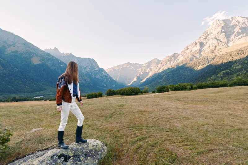 Woman in hiking boots with backpack stands in meadow facing mountains landscape and valley, enjoying nature scenery and panoramic view during outdoor adventure and travel exploration.