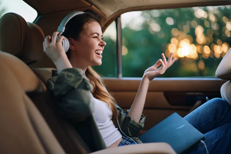 Happy young woman enjoying music in the car, wearing casual clothes, with sunlight and a warm color palette, conveying joy and relaxation during a road trip.