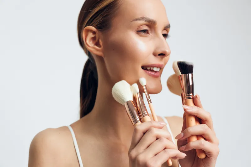 A smiling young woman with clear skin holds several makeup brushes near her face against a plain white background. Concept for beauty, skincare, and cosmetics application.