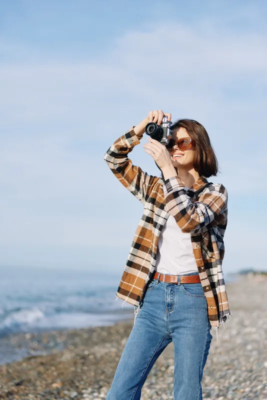 Woman photographer captures a seaside moment with camera, wearing a plaid shirt and jeans, standing on a pebbled beach under blue sky, focused on landscape and memories with soft sunlight.