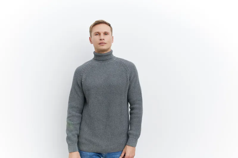 Full body portrait of a man with fair skin, short blonde hair, and blue eyes, wearing a cozy gray turtleneck sweater, standing against a plain white background. Casual fashion look, lifestyle, close-u