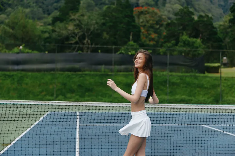 Young athletic woman playing tennis outdoors, wearing a stylish white outfit, exuding happiness on a sunny day, surrounded by lush greenery and a tennis court