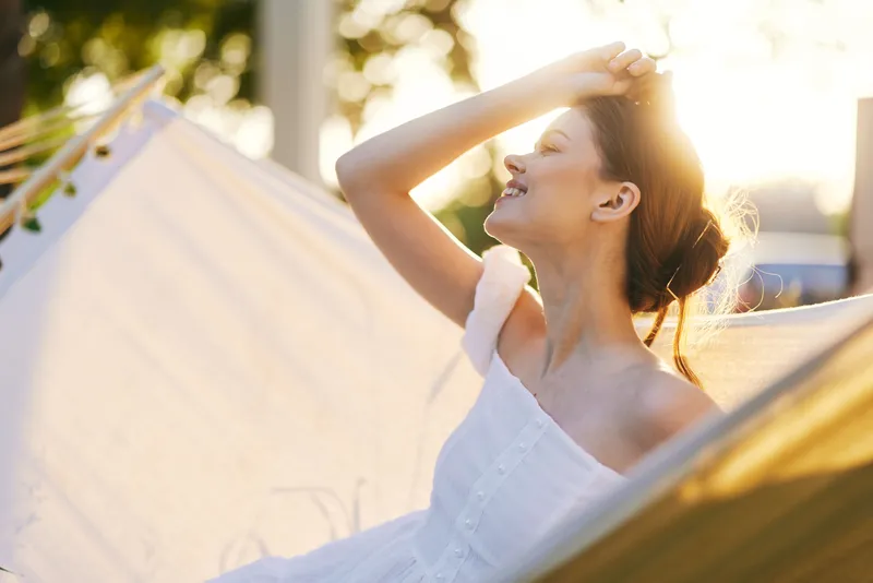 Joyful young woman relaxing in a hammock outdoors, enjoying the sunlight, showcasing a blissful smile and summer vibes in a serene environment