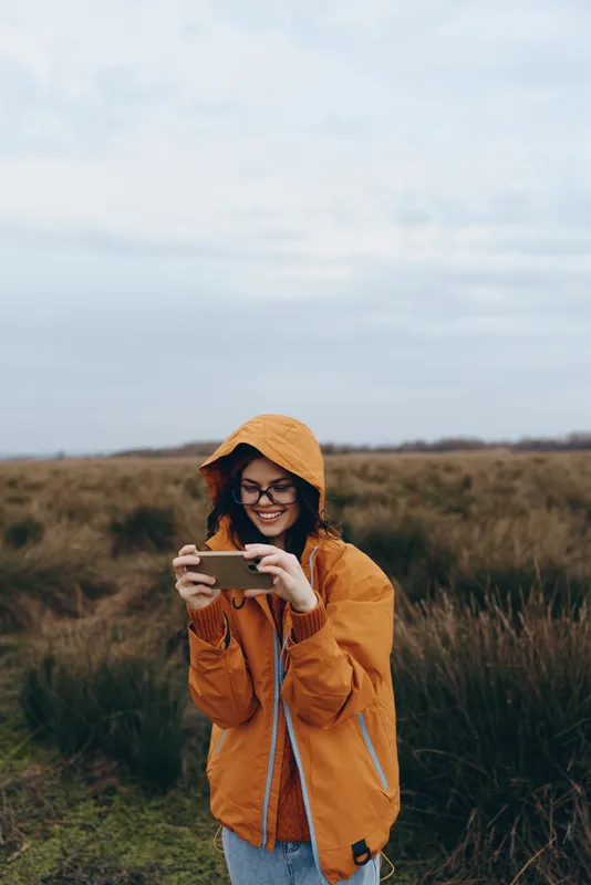 Woman lifestyle outdoor field smartphone jacket smiling glasses cloudy grass, young woman in orange hooded jacket holds a smartphone in a meadow, taking a photo with clouds and daylight.