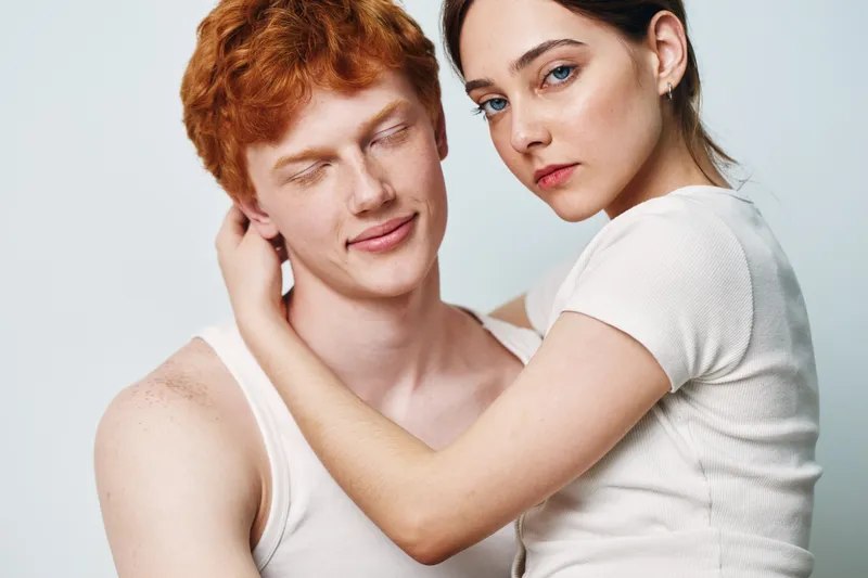 Young couple embracing with gentle expressions on white background. Woman with serious look and man with closed eyes showing affection and closeness in studio photo.