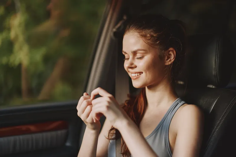 Young woman smiling in a car, wearing a casual tank top, enjoying a moment of relaxation in nature, showcasing happiness and tranquility